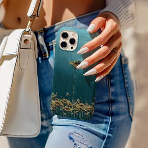Puede incluir: Funda de teléfono de color verde azulado con un diseño floral de girasoles y abejas. La funda tiene un acabado mate y un recorte para la cámara. La funda del teléfono la sostiene una persona que lleva vaqueros azules y un jersey blanco.