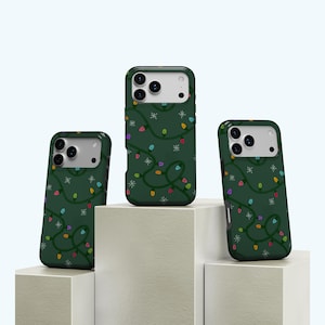 May include: Three green phone cases with a Christmas lights pattern. The cases feature a dark green background with colorful string lights and white snowflakes. The cases are displayed on white geometric blocks.
