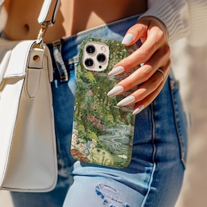 Puede incluir: Una funda de teléfono verde y floral con una rana, agua y flores. La funda la sostiene una persona que lleva vaqueros azules y un jersey blanco. En la imagen se ve un bolso blanco.