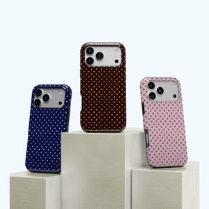 May include: Three phone cases with a polka dot pattern. One case is navy blue with white dots, another is dark brown with white dots, and the third is pink with brown dots. The cases are displayed on white geometric blocks.