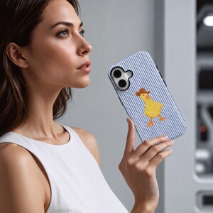May include: A smartphone case with a blue and white striped pattern and a yellow duck wearing a brown cowboy hat. The case has a black camera lens cutout and is held by a person.
