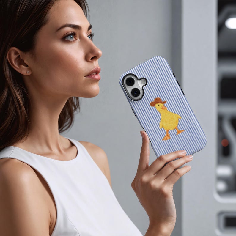 May include: A smartphone case with a blue and white striped pattern and a yellow duck wearing a brown cowboy hat. The case has a black camera lens cutout and is held by a person.