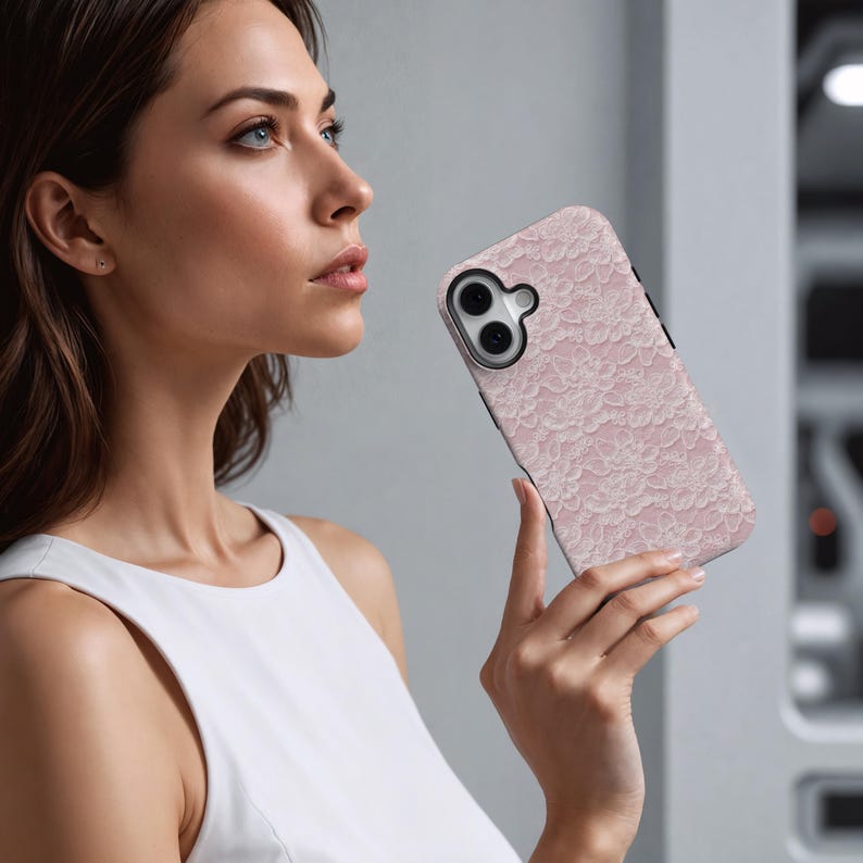 May include: A pink phone case with a delicate lace pattern. The case has a black border and a camera cutout. The phone case is held by a person wearing a white tank top.