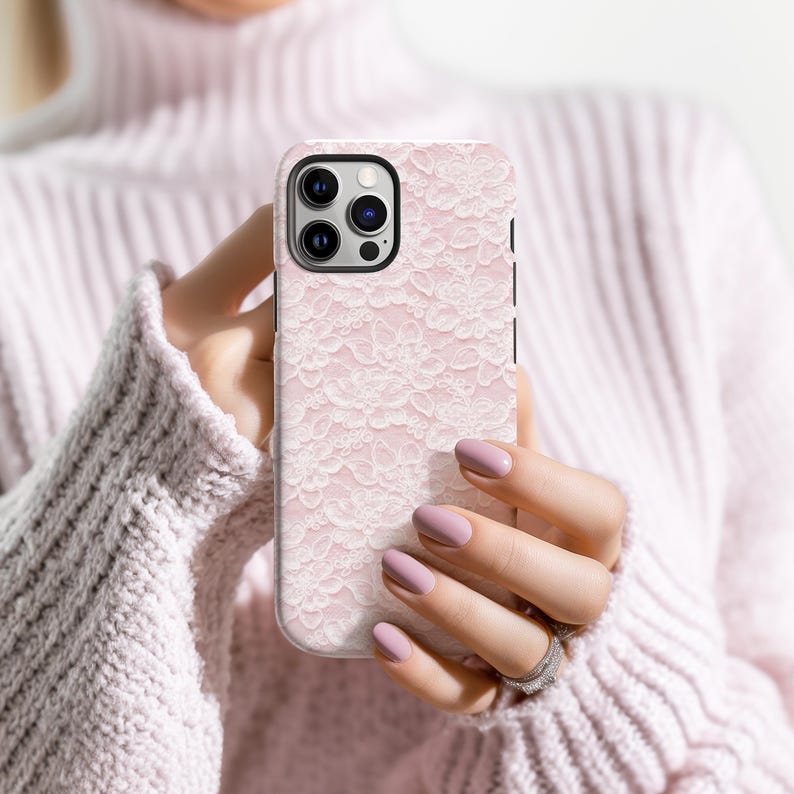 May include: A pink phone case with a white lace pattern. The case has a black camera module and is held by a person wearing a pink sweater. The case is designed to fit a smartphone.