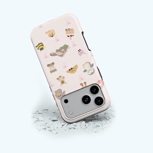 May include: A light pink phone case featuring a repeating pattern of cute cartoon illustrations, including a bee, a croissant, a penguin, and a bear. The case has a black border and a silver camera module.