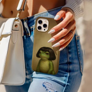 Puede incluir: Una funda de teléfono con una ilustración de una rana verde con un corte de pelo bob negro. La funda tiene un fondo beige y una abertura para la cámara. La funda del teléfono la sostiene una persona que lleva vaqueros azules y un jersey blanco.