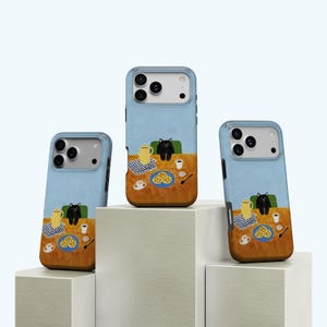 May include: Three phone cases with a whimsical illustration of a black cat at a table. The cases have a light blue background with a brown table, a yellow teapot, and a plate of food. The cat is sitting in a green chair.