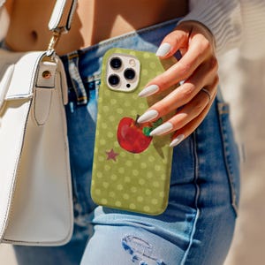 Pode incluir: Uma capa de telefone verde com uma ilustração de maçã vermelha e uma pequena estrela roxa. A capa tem um padrão de bolinhas e é segurada por uma pessoa que usa jeans azuis e uma bolsa branca.