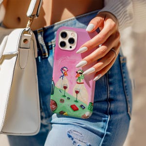 Peut inclure: Coque de téléphone rose avec une illustration fantaisiste de deux oies buvant du vin. Les oies portent des écharpes et des lunettes de soleil, sur une table verte avec des verres à vin et de la nourriture. La coque est tenue par une personne portant un jean bleu.