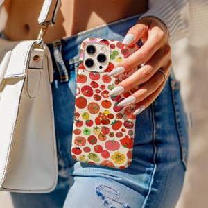 May include: A smartphone case with a colorful tomato pattern. The case features a variety of red, orange, and yellow tomatoes on a cream background. The phone case is held by a person wearing blue jeans.