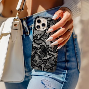 Puede incluir: Funda de teléfono con encaje floral en blanco y negro. La funda presenta un patrón detallado de flores y encaje, con un diseño negro sobre un fondo blanco. La funda del teléfono la sostiene una persona que lleva vaqueros azules.