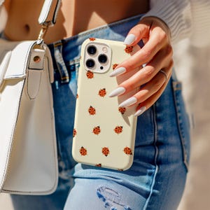 May include: A cream-colored phone case adorned with a pattern of red and black ladybugs. The case is held in front of a person wearing blue jeans. A white purse with a gold strap is visible in the background.