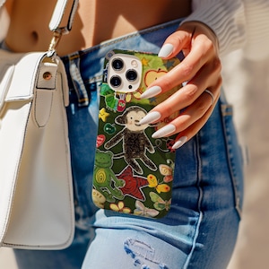 May include: A green phone case with a playful design, including a monkey, frog, butterflies, and stars. The case has a dark green backdrop with colourful illustrations. The phone case is held by a person wearing blue jeans.