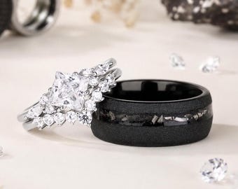 May include: A set of wedding rings. One ring is silver with a square-cut diamond center stone and smaller diamonds. The other ring is black with a textured surface and a band of inlaid stones. The rings are on a white surface.