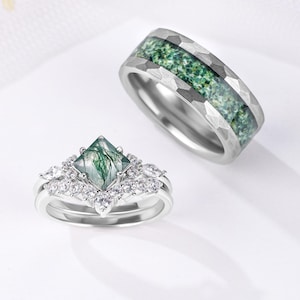 May include: Two rings: a silver engagement ring with a square, green-hued stone and surrounding clear stones, and a silver band with a textured finish and a green stone inlay. The rings are set against a white background.