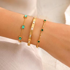 May include: Three gold bracelets are displayed on an arm. One bracelet features square green stones, another has rectangular clear stones, and the third has round green stones. The bracelets are gold-colored.