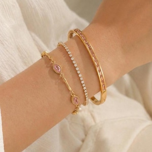 Op de afbeelding: Een collectie gouden armbanden siert een arm. De armbanden omvatten een delicate ketting met roze edelstenen, een dunne band met kleine heldere stenen en een bredere band met rechthoekige roze stenen. Witte achtergrond.