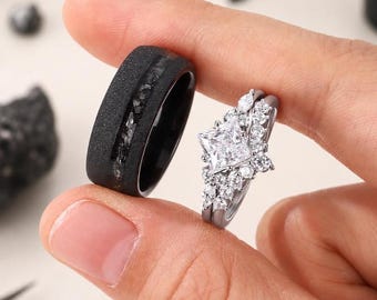 Puede incluir: Un anillo negro con superficie texturizada e incrustación oscura, junto a un anillo de compromiso plateado con una piedra central cuadrada y piedras más pequeñas alrededor. Los anillos se sostienen sobre un fondo neutro.