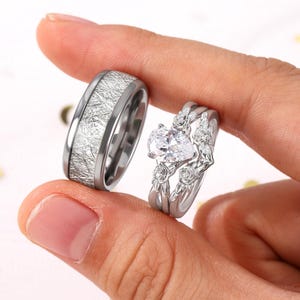 May include: Two rings are displayed. One is a silver band with a textured, light-colored inlay. The other is a silver engagement ring with a pear-shaped, clear gemstone and floral detailing. The rings are held between fingers.