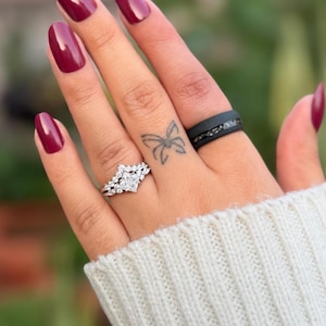 Puede incluir: Una mano con esmalte de u&ntilde;as burdeos y un tatuaje de mariposa. La mano luce dos anillos: un anillo de compromiso plateado con una piedra cuadrada central y un anillo de banda negro con piedras incrustadas.
