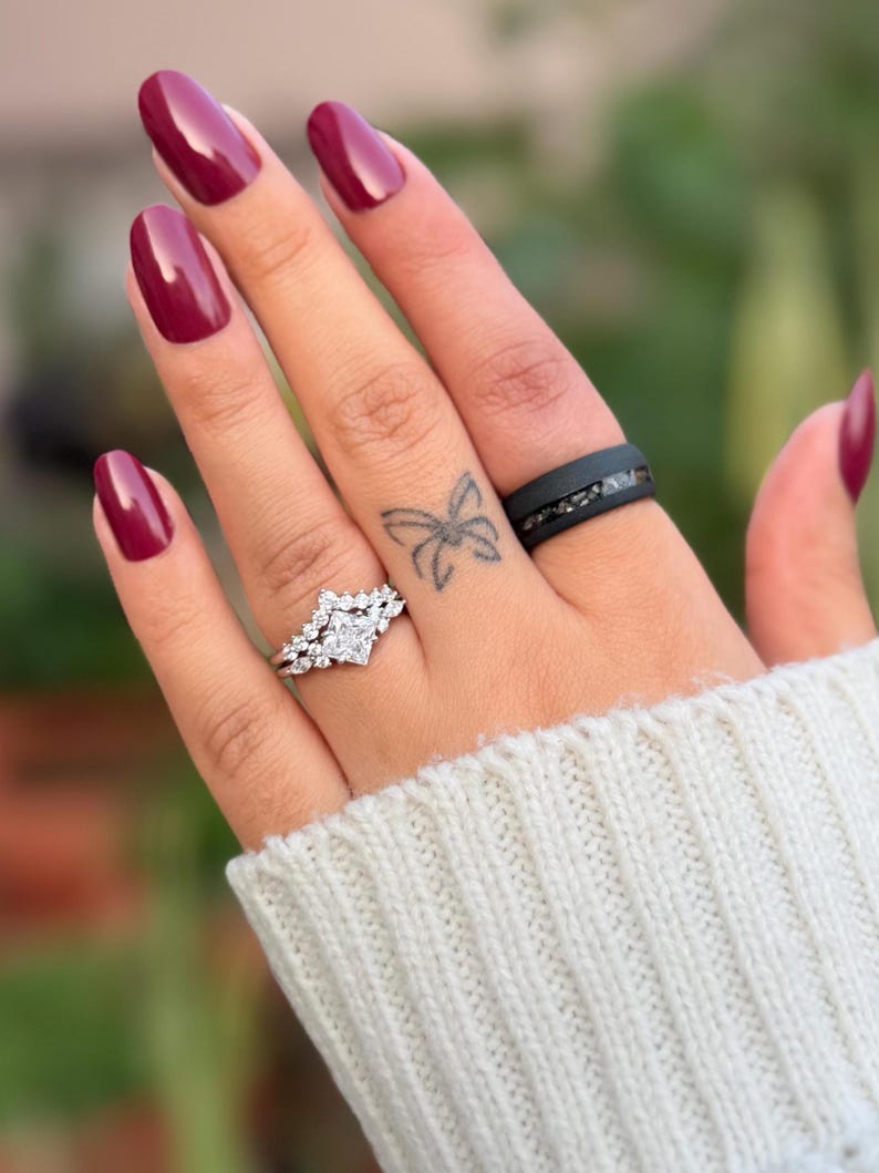 Puede incluir: Una mano con esmalte de u&ntilde;as burdeos y un tatuaje de mariposa. La mano luce dos anillos: un anillo de compromiso plateado con una piedra cuadrada central y un anillo de banda negro con piedras incrustadas.