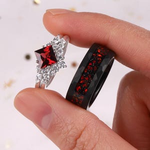 Puede incluir: Un anillo de compromiso plateado con una piedra preciosa roja cuadrada y piedras blancas más pequeñas. Un anillo negro con motas rojas está al lado. Los anillos están sostenidos por los dedos de una persona.