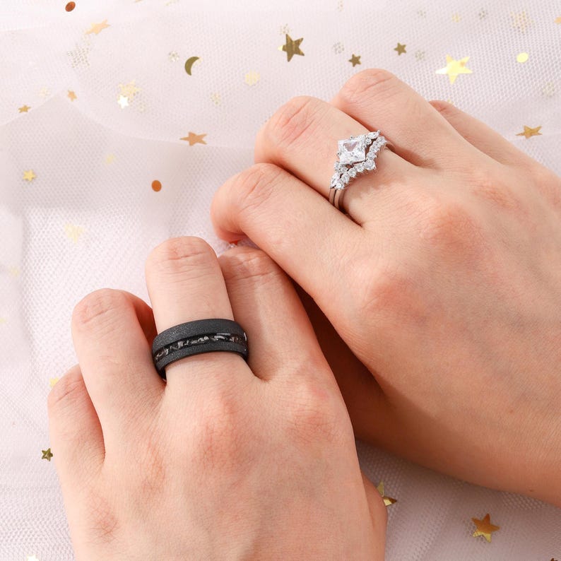 Puede incluir: Se muestran dos manos, cada una con un anillo. Una mano tiene un anillo negro con una banda texturizada. La otra mano tiene un anillo plateado con una piedra cuadrada y transparente y piedras m&aacute;s peque&ntilde;as alrededor. El fondo es una tela rosa claro con estrellas doradas.