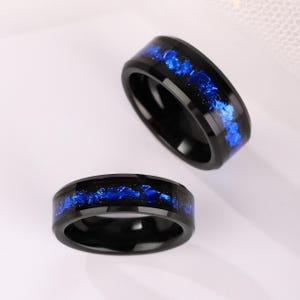 Könnte beinhalten: Zwei schwarze Ringe mit blauem Inlay. Die Ringe haben eine polierte, reflektierende Oberfläche. Das blaue Inlay hat ein strukturiertes, fragmentiertes Aussehen und bildet einen auffälligen Kontrast zum schwarzen Band. Die Ringe werden auf einer weißen Oberfläche präsentiert.