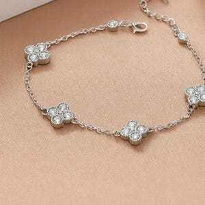 Op de afbeelding: Een zilveren armband met klavermotieven met vier bladeren, elk bezet met heldere, sprankelende edelstenen. De armband heeft een fijne ketting en een veilige sluiting. De sieraden worden gepresenteerd op een zacht, perzikroze oppervlak.