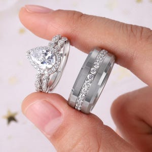 May include: Two silver rings. One has a pear-shaped gemstone with surrounding smaller stones. The other is a wider band with a row of small, clear stones. Both rings are held up by a person's fingers.