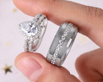 May include: A pair of silver rings. One ring features a pear-shaped gemstone surrounded by smaller stones. The other ring is a wider band with a row of small, clear stones. Both rings are held up by a person's fingers.