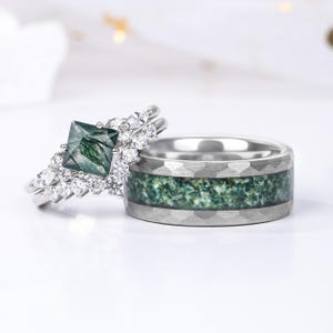 Op de afbeelding: Twee trouwringen. Eén ring heeft een vierkante, groene edelsteen omringd door kleinere heldere stenen. De andere ring is zilverkleurig met een getextureerde band en een groene, gespikkelde inleg. De ringen staan tegen een witte achtergrond.