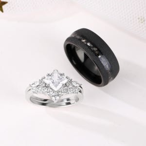 Puede incluir: Dos anillos de boda. Uno es una banda plateada con una piedra central de corte cuadrado y piedras m&aacute;s peque&ntilde;as alrededor. El otro es una banda negra con una superficie texturizada y una incrustaci&oacute;n m&aacute;s clara.