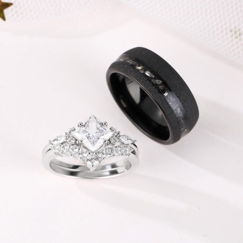 Puede incluir: Dos anillos de boda. Uno es una banda plateada con una piedra central de corte cuadrado y piedras m&aacute;s peque&ntilde;as alrededor. El otro es una banda negra con una superficie texturizada y una incrustaci&oacute;n m&aacute;s clara.