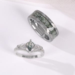 Könnte beinhalten: Ein silberfarbenes Ring-Set bestehend aus einem Band mit facettiertem Rand und Moosachat-Einlage sowie einem V-förmigen Ring mit einem drachenförmigen Moosachat-Mittelstein, flankiert von kleinen klaren Steinen auf einem Pavé-Band.