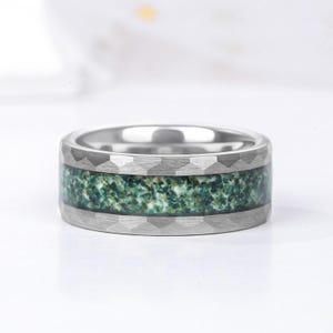 Op de afbeelding: Een zilveren ring met een getextureerde band en een centrale inleg van groen en wit gespikkeld materiaal. De ring heeft een brede band en een gepolijst interieur.