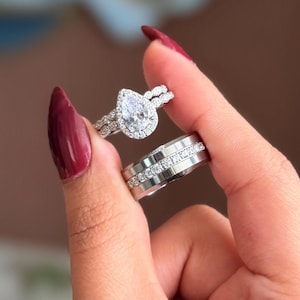 Può includere: Due anelli d'argento, uno con un diamante a forma di pera circondato da pietre più piccole, e l'altro con una fascia di piccole pietre trasparenti. Gli anelli sono presentati su uno sfondo sfocato.