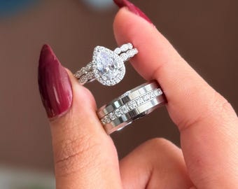 May include: A pair of silver rings, one with a pear-shaped diamond surrounded by smaller stones, and another with a band of small, clear stones. The rings are held up against a blurred background.