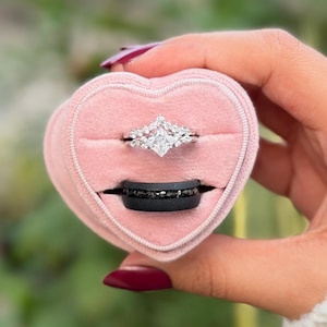 Puede incluir: Una caja de anillos rosa en forma de coraz&oacute;n con dos anillos. El anillo superior es plateado con un diamante cuadrado central y piedras m&aacute;s peque&ntilde;as. El anillo inferior es negro con piedras oscuras incrustadas.
