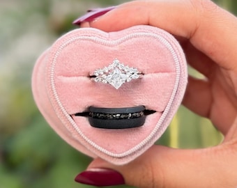 May include: A pink heart-shaped ring box holding two rings. The top ring is silver with a central square diamond and smaller surrounding stones. The bottom ring is black with embedded dark stones. The box is held by a person.