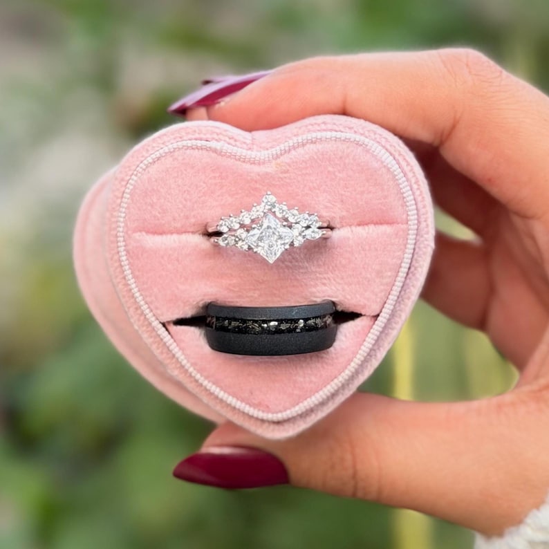 Puede incluir: Una caja de anillos rosa en forma de coraz&oacute;n con dos anillos. El anillo superior es plateado con un diamante cuadrado central y piedras m&aacute;s peque&ntilde;as. El anillo inferior es negro con piedras oscuras incrustadas.