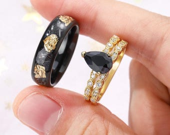 Op de afbeelding: Twee ringen: een zwarte ring met goudbladaccenten en een gouden ring bezet met een zwarte druppelvormige steen en kleine heldere stenen. De ringen worden tussen de vingers gehouden.