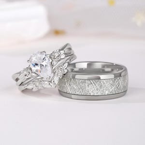 Pode incluir: Um conjunto de dois anéis de prata. Um anel apresenta uma gema clara em forma de pêra e detalhes florais. O outro anel é uma faixa larga com um padrão texturizado entrecruzado. Ambos os anéis em um fundo branco.