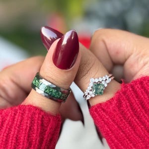 Könnte beinhalten: Zwei Ringe werden gezeigt. Ein Ring hat ein gehämmertes Silberband mit einem grünen Steineinsatz. Der andere Ring hat ein Silberband mit einem quadratischen grünen Stein und umliegenden weißen Steinen. Der Hintergrund ist verschwommen, mit einem roten Pullover und burgunderfarbenen Nägeln.