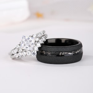 Puede incluir: Un conjunto nupcial con un anillo de compromiso plateado con una piedra central de corte cuadrado y diamantes de acento circundantes. Tambi&eacute;n se muestra una alianza de boda negra con un acabado texturizado e incrustaciones.