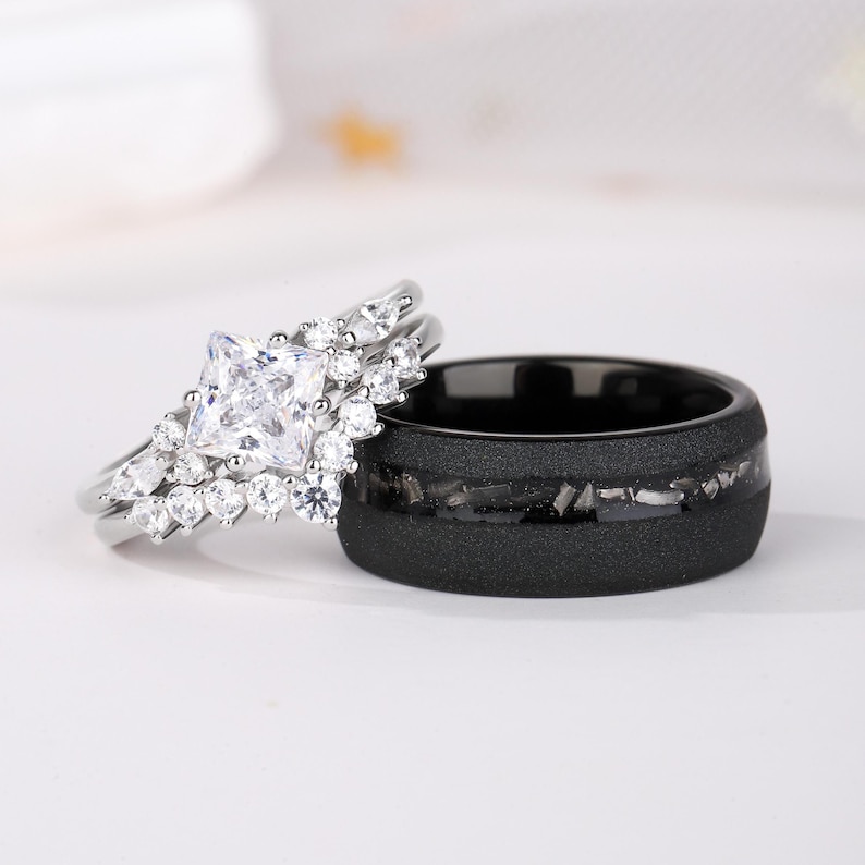 Puede incluir: Un conjunto nupcial con un anillo de compromiso plateado con una piedra central de corte cuadrado y diamantes de acento circundantes. Tambi&eacute;n se muestra una alianza de boda negra con un acabado texturizado e incrustaciones.
