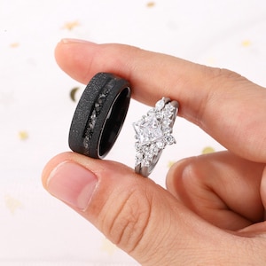 Puede incluir: Un anillo negro con una superficie texturizada y una banda de piedras de color gris oscuro junto a un juego de anillos plateados con un diamante de talla cuadrada y piedras de acento m&aacute;s peque&ntilde;as. Los anillos son sostenidos por una mano.