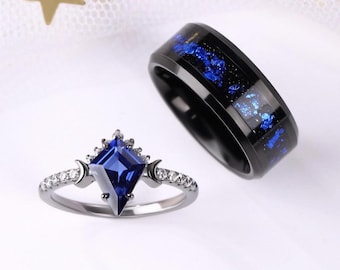Op de afbeelding: Twee ringen: een zilveren ring met een vierkante, donkerblauwe edelsteen en een zwarte ring met blauwe vlekken. De zilveren ring heeft een rij kleine, heldere stenen en halve maan details. De zwarte ring heeft een band van blauw en zwart materiaal.