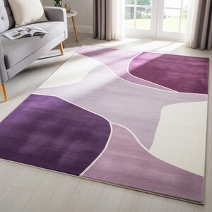Könnte beinhalten: Ein rechteckiger Teppich mit einem abstrakten Design in Lila- und Weißtönen. Der Teppich hat ein geometrisches Muster mit geschwungenen Linien und verschiedenen Lilatönen, von tiefem Violett bis zu hellem Lavendel. Der Teppich liegt auf einem hellen Holzboden.