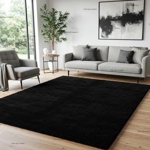 May include: A large, rectangular black shag rug is the focal point in a living room. The rug's deep pile contrasts with the light wooden floor. A grey sofa and armchair, along with a modern painting, complete the room's decor.